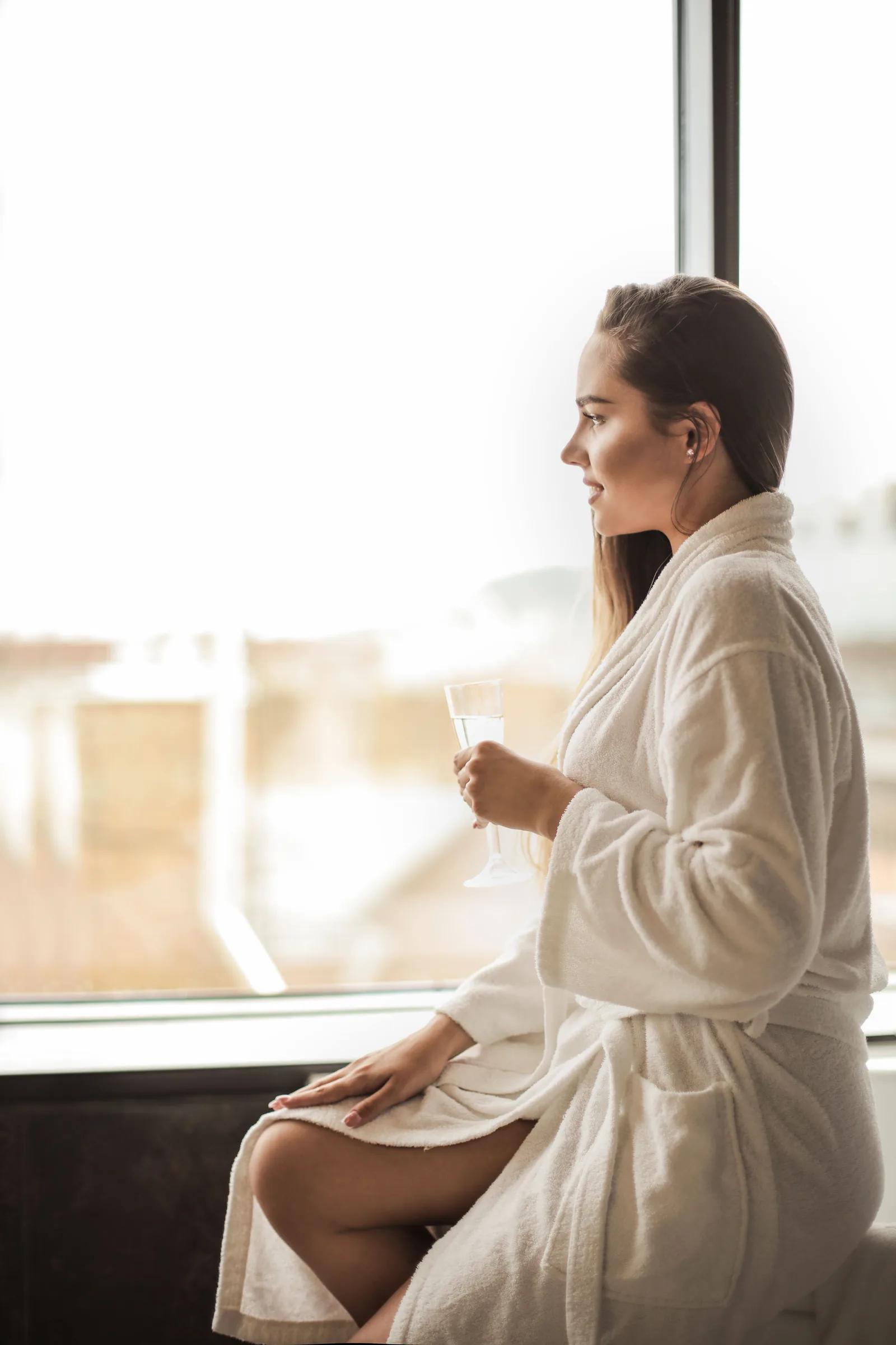 A woman in a white robe sits by a tall window in morning light