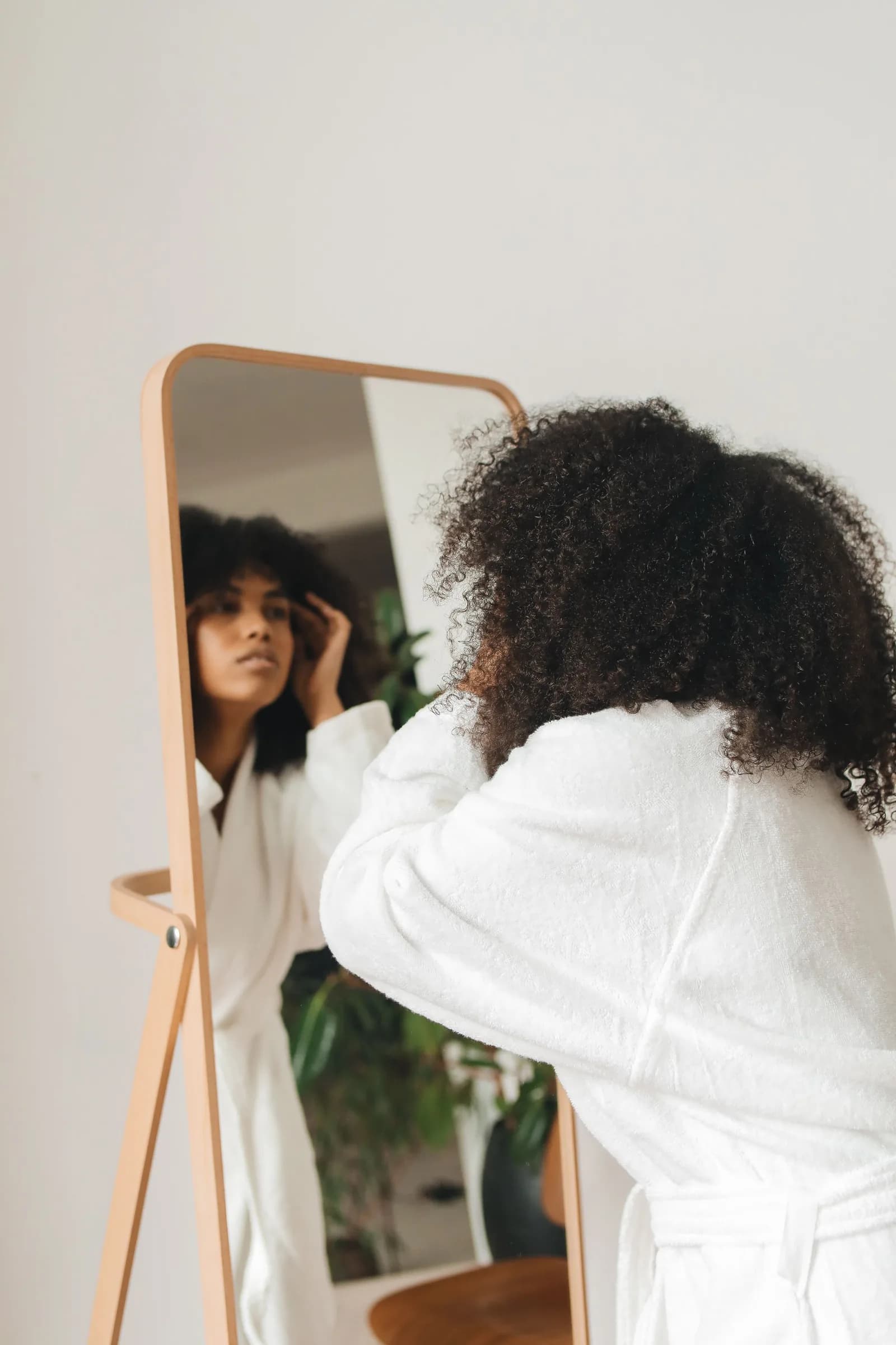 A woman in a white robe looks into a standing mirror during her morning routine