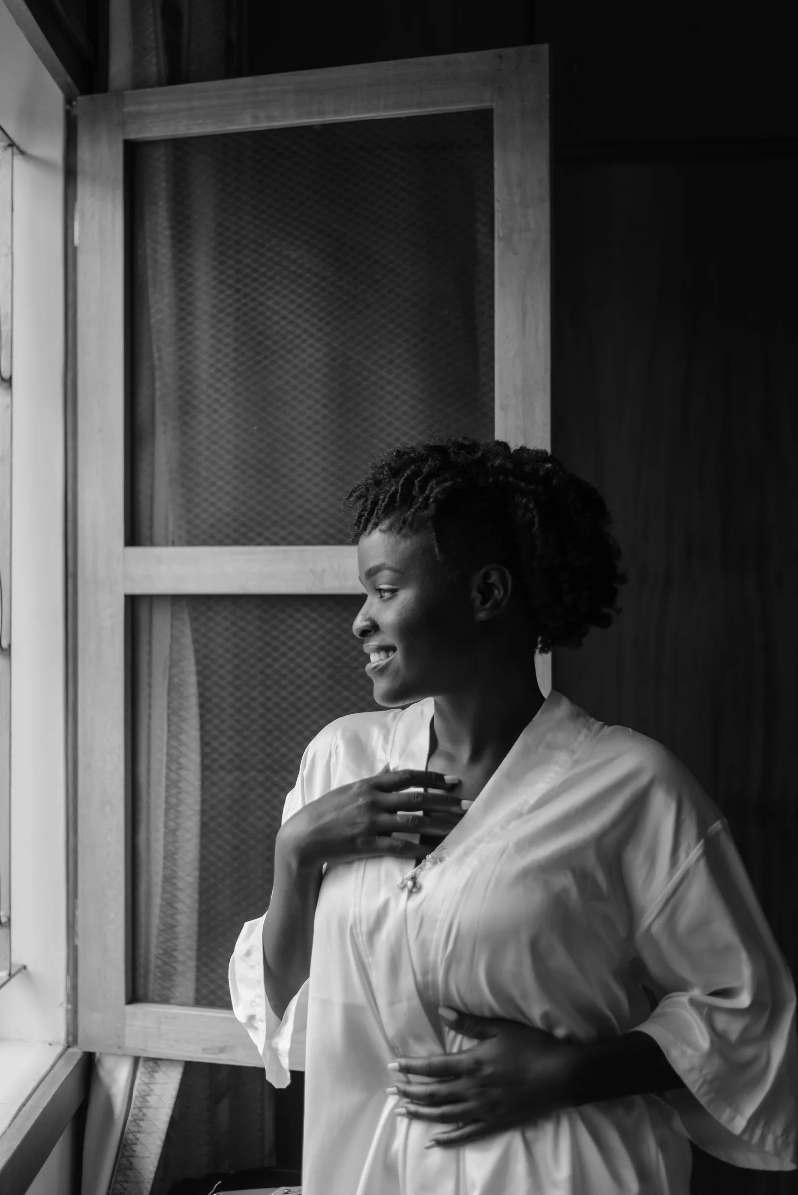 A smiling woman in a light robe looks out of an open window