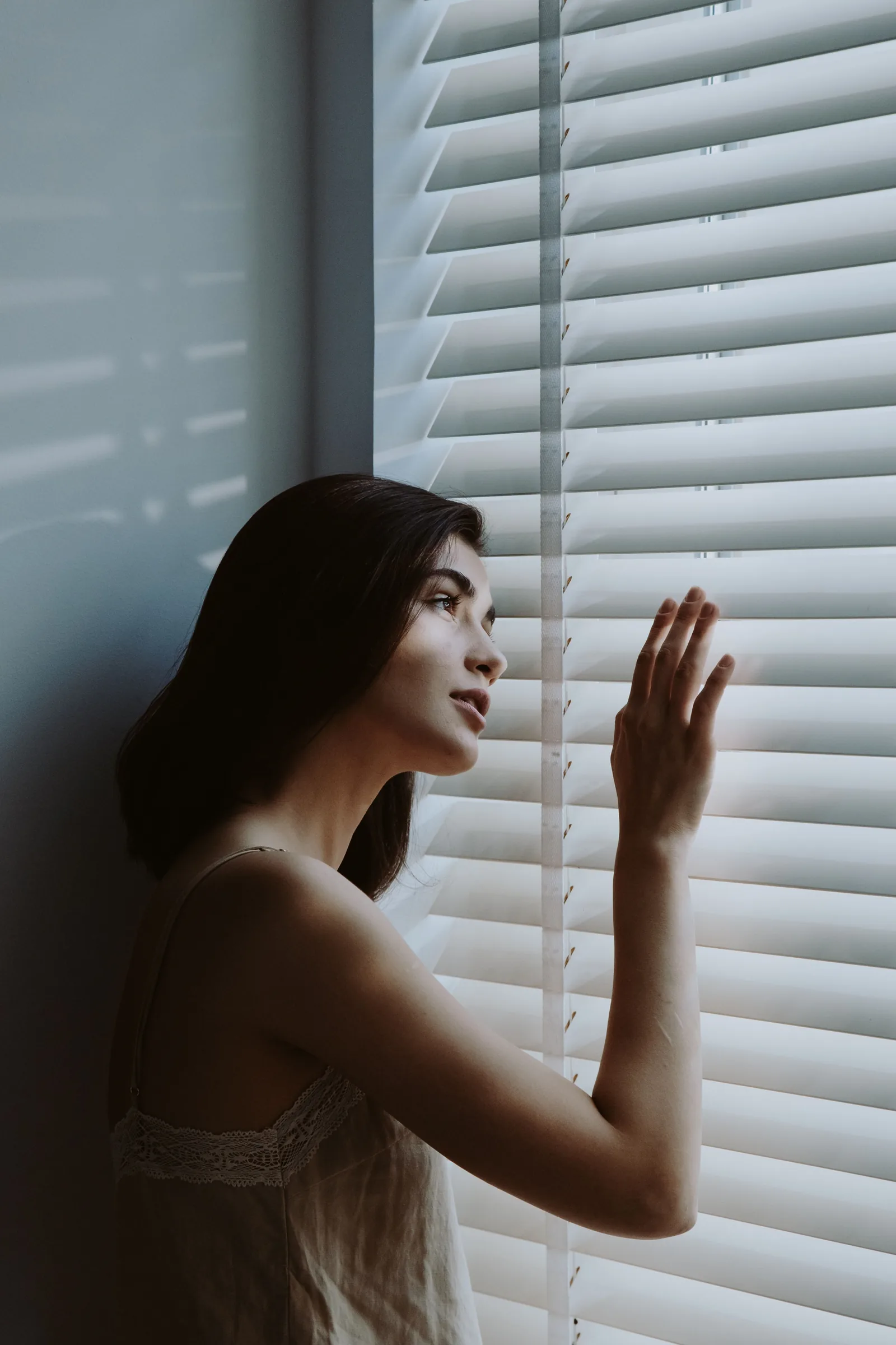 A woman in a delicate silk slip looks through window blinds in soft evening light